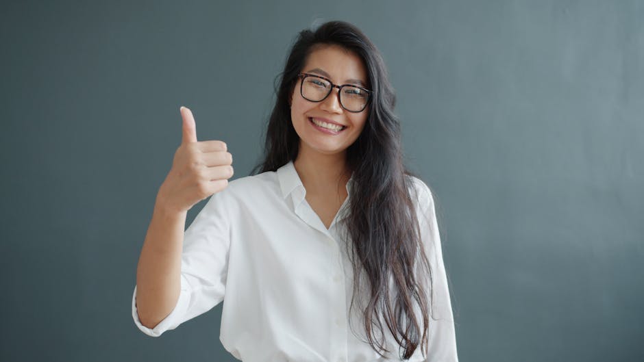 Smiling woman in glasses and white shirt giving a thumbs up against gray background.